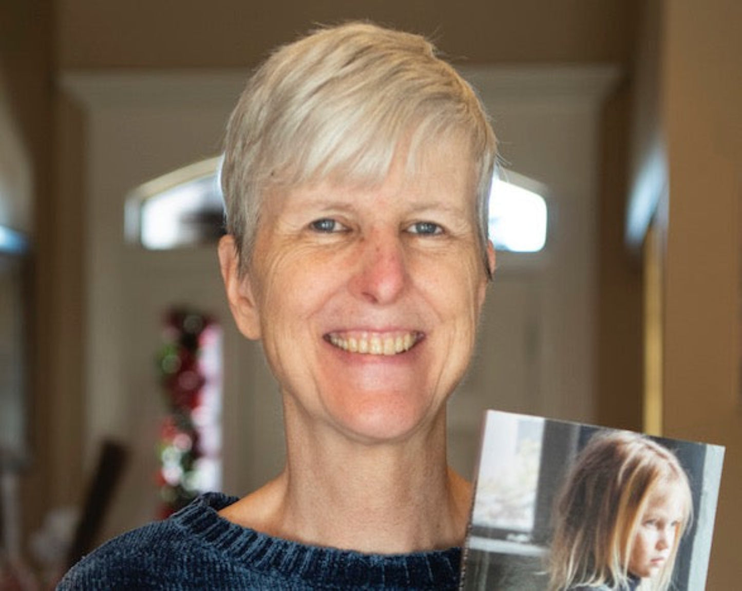 De holding a copy of "Born a Poor, Black, Indian, White Girl", her memoir. Photo shows a poor girl inside a house looking out the window, like she's dreaming of a better life.